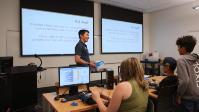 An instructor in front of projector screens holding a robotics kit, teaching teenagers.